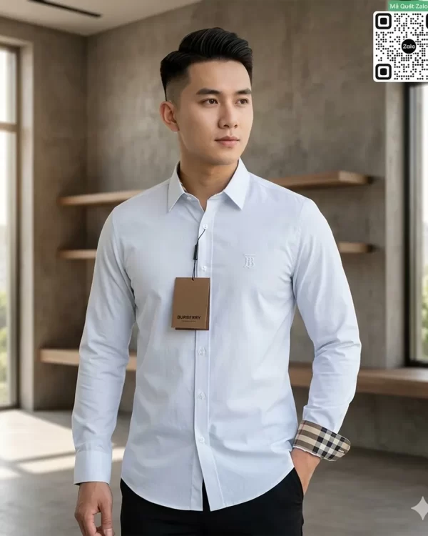 Người mẫu nam với kiểu tóc undercut nam tính đang diện áo sơ mi dài tay màu trắng của Burberry phối cùng quần tây đen, tay áo bên trái xắn nhẹ lộ viền kẻ sọc, ngực áo đính tag giấy nâu sang trọng.