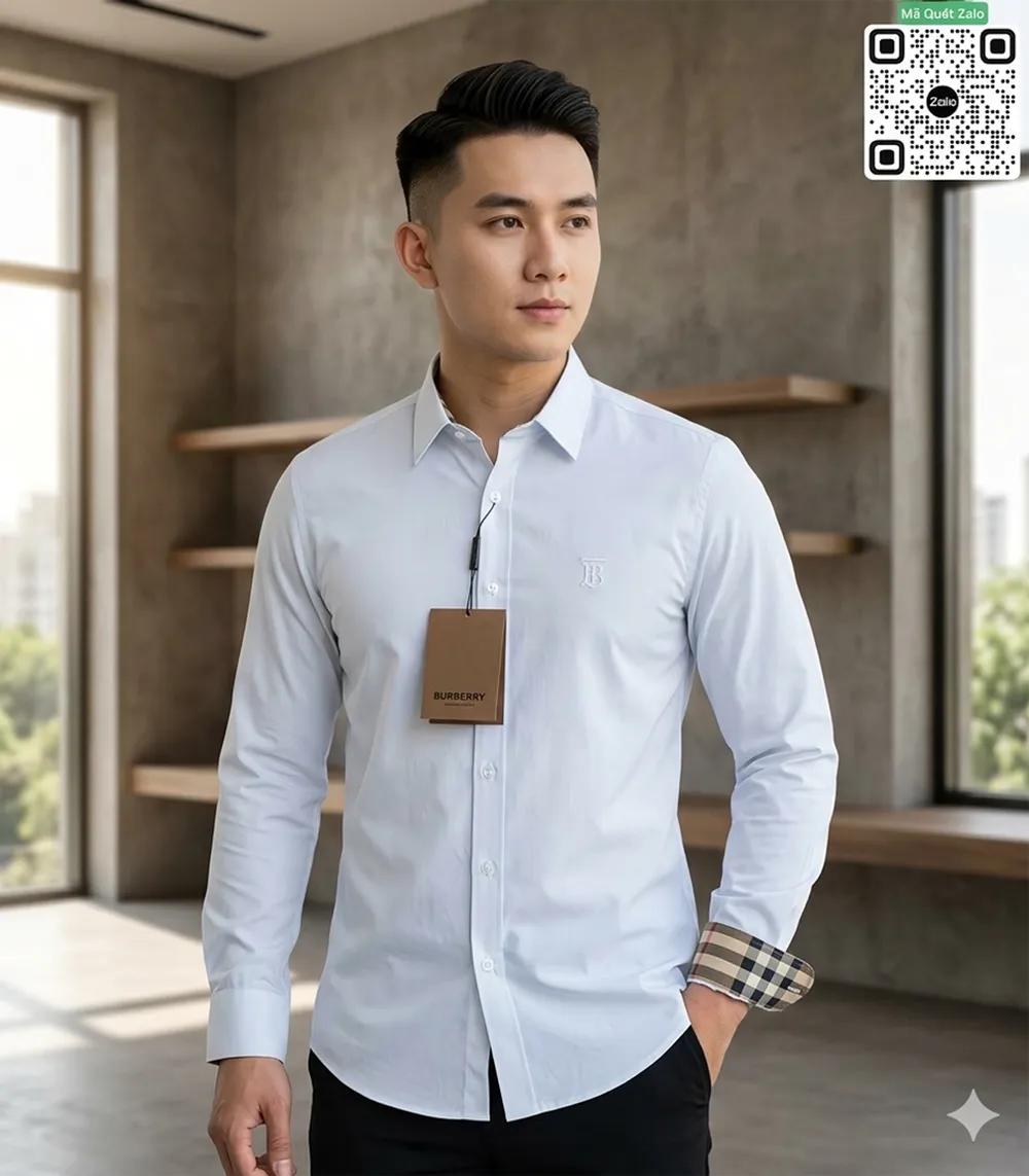 Người mẫu nam với kiểu tóc undercut nam tính đang diện áo sơ mi dài tay màu trắng của Burberry phối cùng quần tây đen, tay áo bên trái xắn nhẹ lộ viền kẻ sọc, ngực áo đính tag giấy nâu sang trọng.