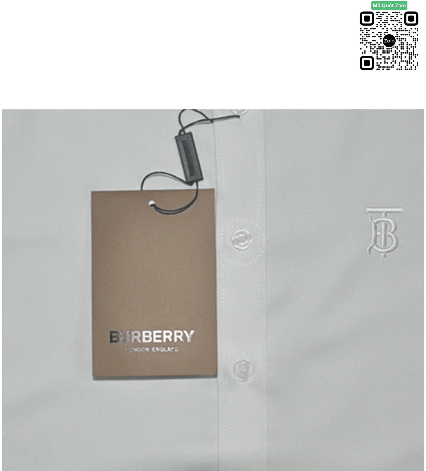 Ảnh chụp sát ngực áo sơ mi trắng hiển thị logo chữ lồng TB thêu nổi bằng chỉ trắng tinh xảo, bên cạnh là sợi dây treo chiếc tag mác bằng giấy thô màu nâu in chữ Burberry vàng ánh kim sang trọng.